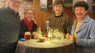 Vier Menschen sitzen um einen Tisch herum und lächeln. Zwei Frauen und ein Mann, wobei eine Frau ein Bierglas hält. Flaschen und Gläser stehen auf dem Tisch. Dahinter eine Holzwand mit gerahmten Bildern.