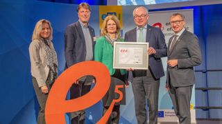 Fünf Personen stehen zusammen, möglicherweise bei einer Preisverleihung. Die Frau in der Mitte hält eine gerahmte Urkunde mit dem Text 'eee'. Die beiden Männer auf beiden Seiten lächeln und schauen zu ihr. Hinter ihnen befindet sich eine Wand mit einer großen Zahl '5'.