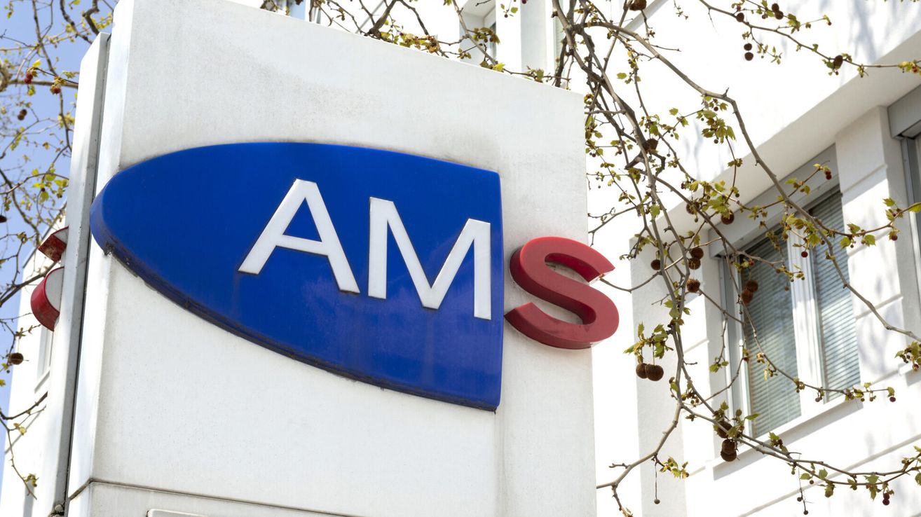 Ein blaues und weißes Schild mit den Buchstaben AMS in Weiß auf einer weißen Gebäudewand, mit einem roten S daneben und einem Zweig mit Blättern darüber.