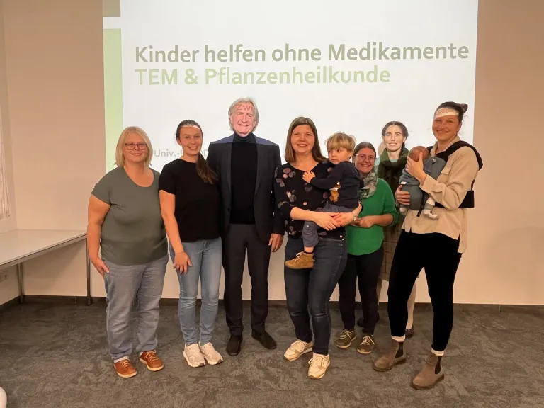 Eine Gruppe von Menschen steht vor einer weißen Leinwand. Die Frau rechts hält ein Baby. Ein Mann in der Mitte trägt eine Maske. Der Text auf der Leinwand lautet 'Kinder helfen ohne Medikamente TEM & Pflanzenheilkunde'.