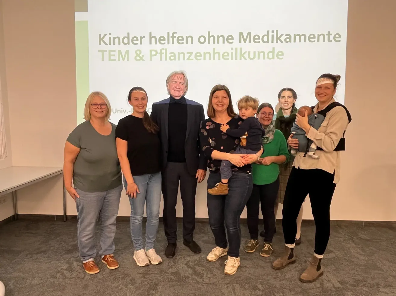 Eine Gruppe von Menschen steht vor einer weißen Leinwand. Die Frau rechts hält ein Baby. Ein Mann in der Mitte trägt eine Maske. Der Text auf der Leinwand lautet 'Kinder helfen ohne Medikamente TEM & Pflanzenheilkunde'.