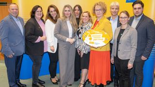 Eine Gruppe von Menschen steht in einem Raum und lächelt. Eine Frau hält ein gelbes Schild mit dem Text 'Gemeinsam gegen Gewalt'. Sie tragen alle formelle Kleidung. Der Raum hat eine gelbe Wand mit blauen Streifen und einem weißen Boden.