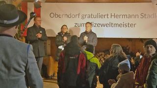 Drei Personen stehen vor einem Banner, auf dem 'Oberndorf gratuliert Hermann zum izeweltmeister' steht. Sie sprechen in Mikrofone. Vor ihnen stehen einige Leute. Eine Person steht auf der linken Seite.