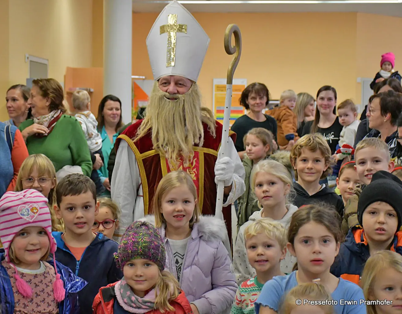 Eine Gruppe von Kindern und Erwachsenen versammelt sich um einen als Bischof verkleideten Mann. Der Mann hält einen Stab mit einem goldenen Kreuz.