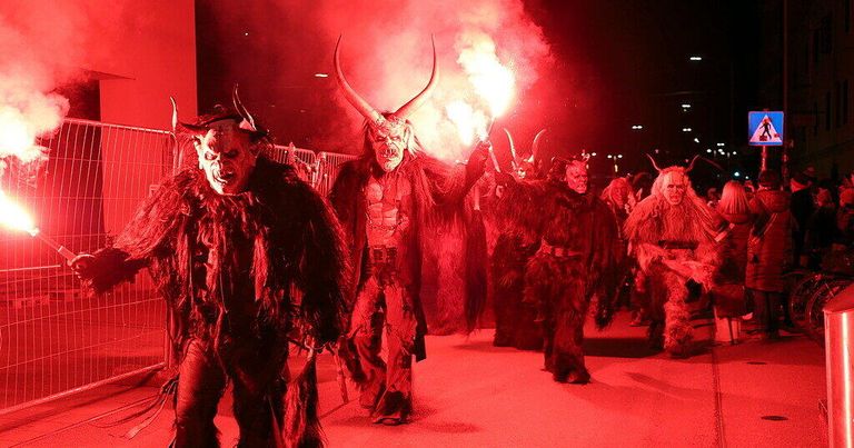 Eine Gruppe von Menschen in Hörnermasken geht unter roter Beleuchtung. Sie halten brennende Fackeln. Der Boden ist rot. Hinter ihnen beleuchtet ein rotes Licht den Bereich.