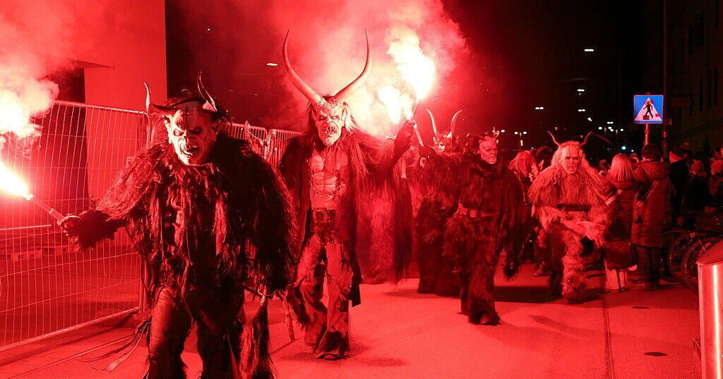 Eine Gruppe von Menschen in Hörnermasken geht unter roter Beleuchtung. Sie halten brennende Fackeln. Der Boden ist rot. Hinter ihnen beleuchtet ein rotes Licht den Bereich.