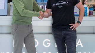 Zwei Männer schütteln sich die Hände vor einem weißen Tresen mit einem großen 'OGG'-Logo. Einer trägt einen grünen Pullover, der andere ein schwarzes T-Shirt. Hinter ihnen steht eine Frau in der Nähe eines Regals mit verschiedenen Produkten.