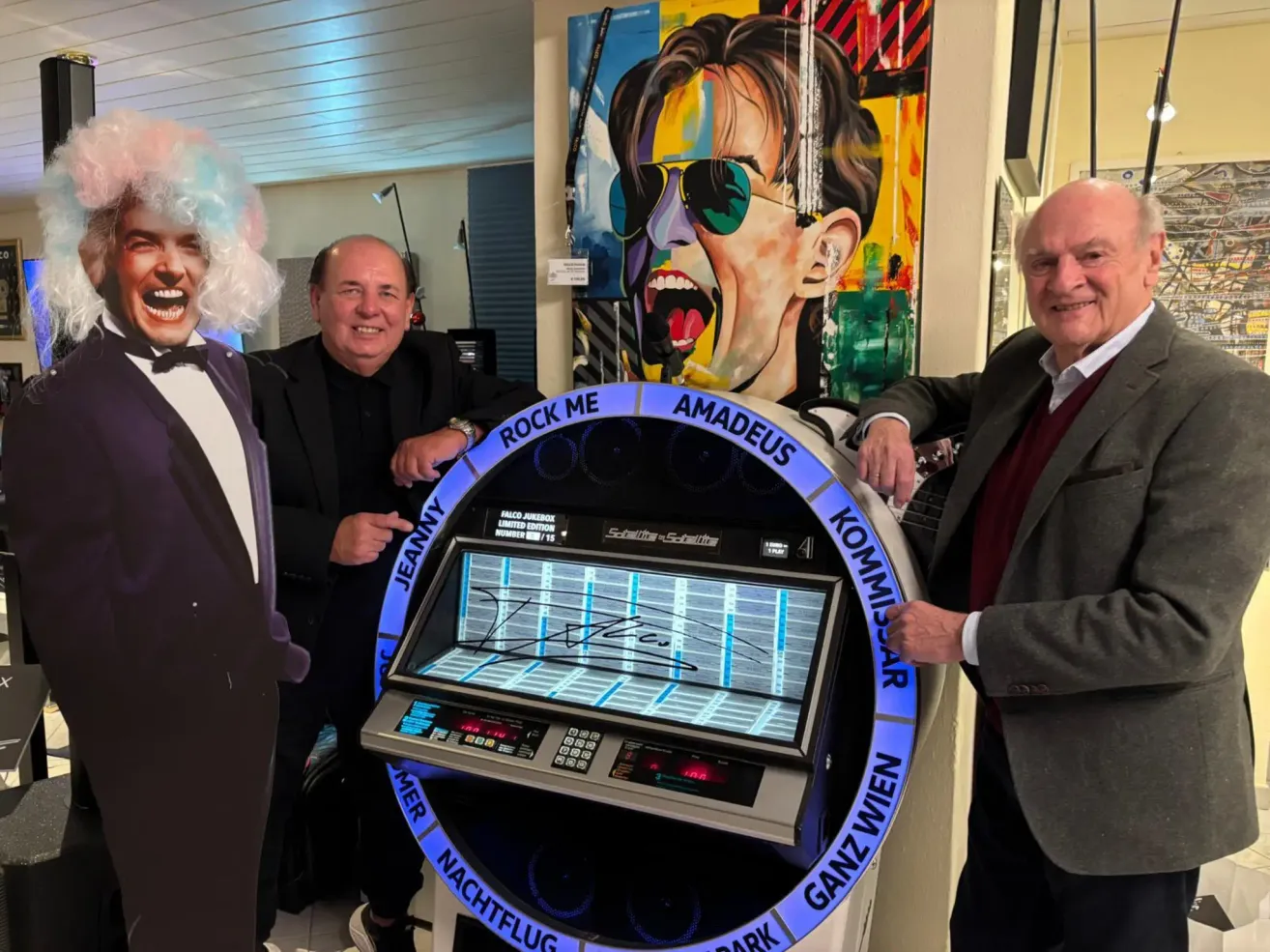 Drei Männer posieren neben einem alten Jukebox, auf dem ein Schild mit den Worten Rock Me Amadeus steht. Der Mann auf der linken Seite trägt eine Perücke. Hinter ihnen ist ein farbenfrohes Bild eines Menschen mit Sonnenbrille.