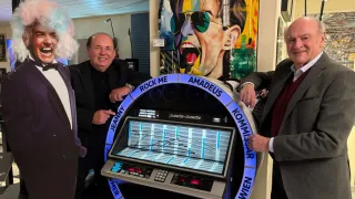Drei Männer posieren neben einem alten Jukebox, auf dem ein Schild mit den Worten Rock Me Amadeus steht. Der Mann auf der linken Seite trägt eine Perücke. Hinter ihnen ist ein farbenfrohes Bild eines Menschen mit Sonnenbrille.