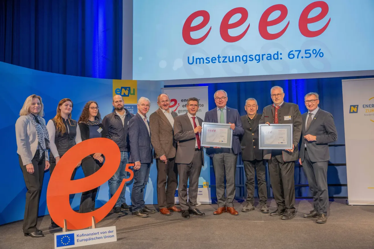 Eine Gruppe von Männern und Frauen auf der Bühne, die Urkunden halten. Sie sind alle formell gekleidet. Hinter ihnen ist ein großes Banner mit rotem Text.