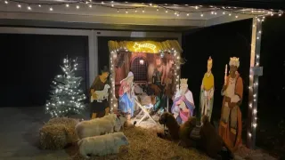 Eine Krippe wird nachts vor einem Haus ausgestellt und zeigt Maria, Josef und das Jesuskind mit Tieren und Weisen, beleuchtet durch Lichterketten und einen Weihnachtsbaum.