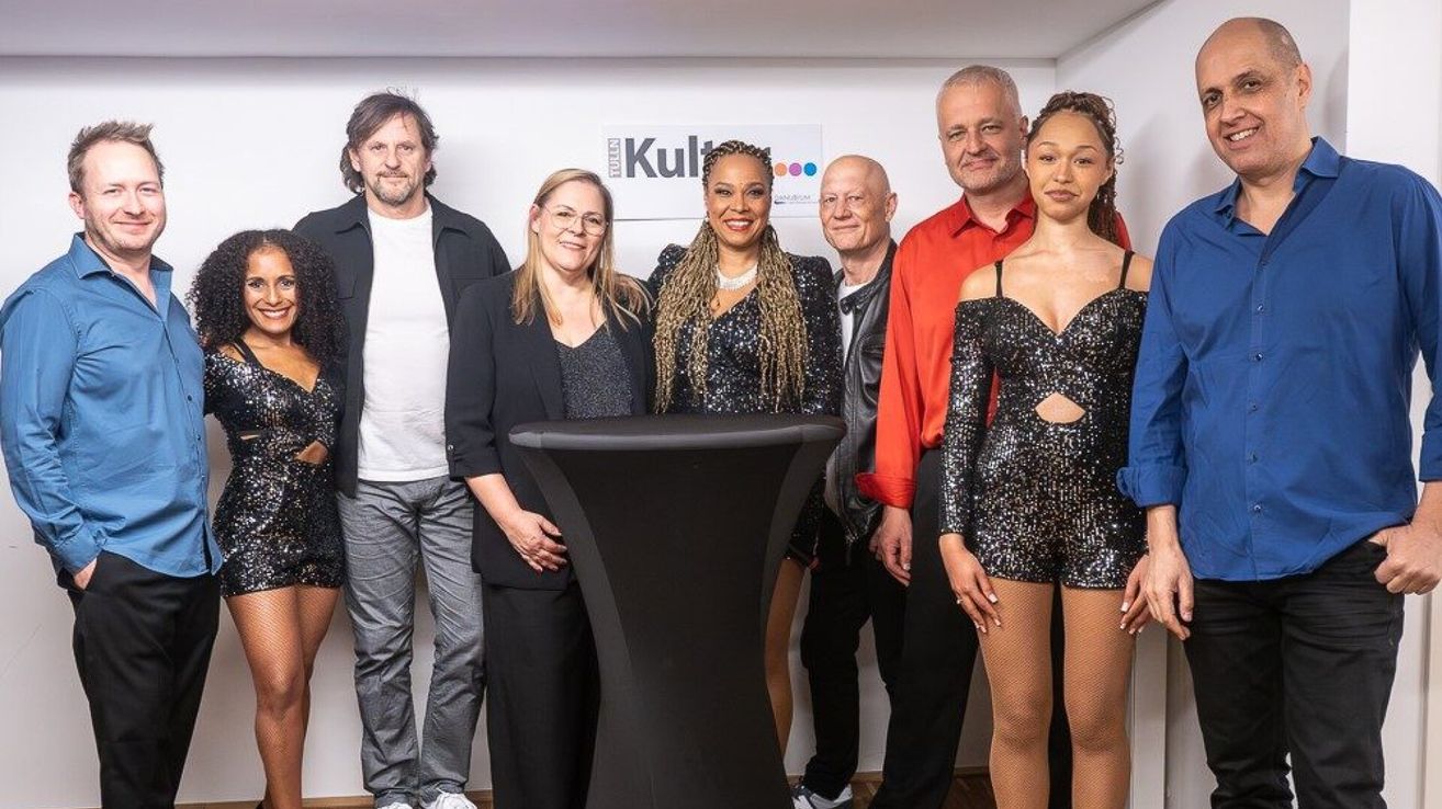 Eine Gruppe von Menschen, darunter zwei Frauen und vier Männer, posiert für ein Foto in einem Raum mit einer weißen Wand. Eine Frau trägt ein funkelndes Kleid, und ein Mann hinter einem Schreibtisch lächelt.
