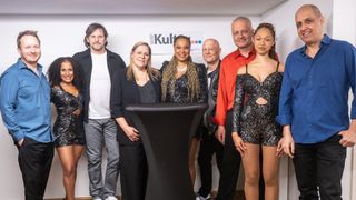 Eine Gruppe von Menschen, darunter zwei Frauen und vier Männer, posiert für ein Foto in einem Raum mit einer weißen Wand. Eine Frau trägt ein funkelndes Kleid, und ein Mann hinter einem Schreibtisch lächelt.