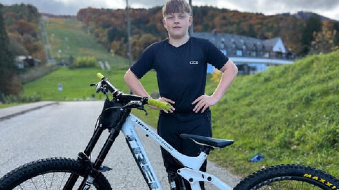 Ein junger Junge steht neben einem Mountainbike mit den Händen auf den Hüften, trägt ein schwarzes T-Shirt und Hose und lächelt leicht. Dahinter befindet sich eine Rasenfläche mit Bäumen und einem Gebäude.