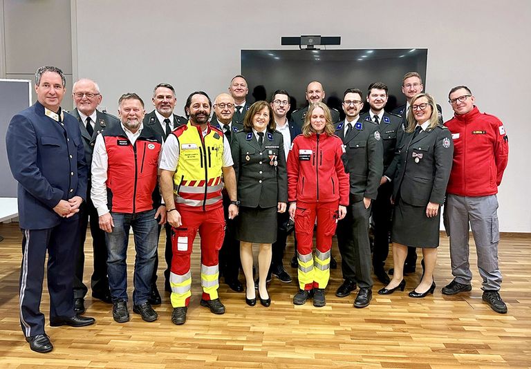 Eine Gruppe von Personen in Uniformen und Schutzausrüstungen posiert für ein Foto in einem Konferenzraum. Die Frau in der Mitte trägt eine rote Jacke.