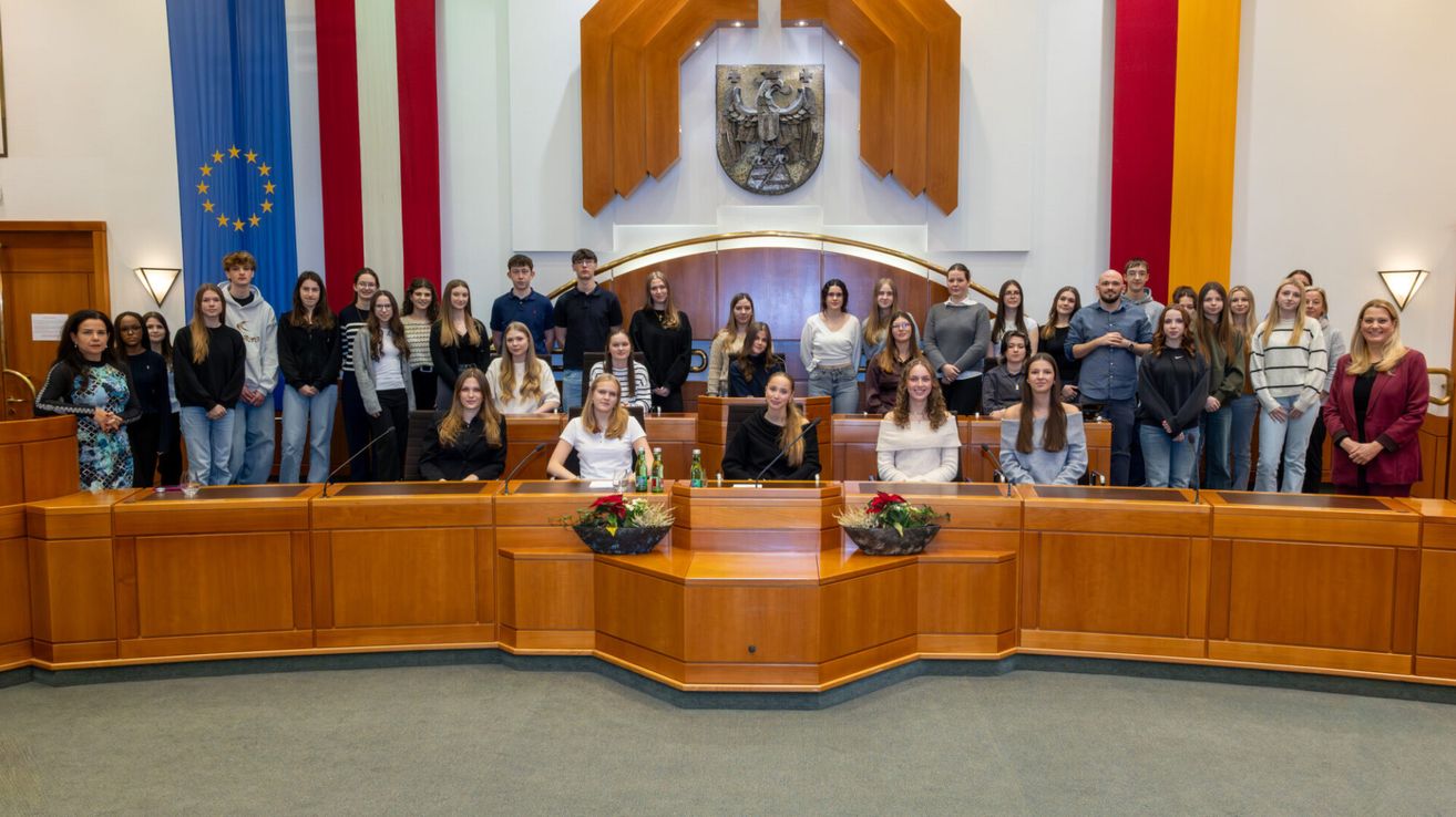 Eine Gruppe von Personen in formeller Kleidung posiert für ein Foto in einem Konferenzraum mit Holztischen und Mikrofonen.