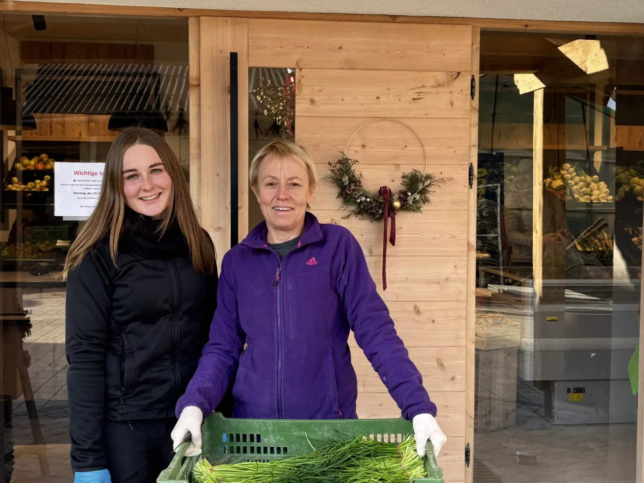 Zwei Frauen stehen vor einem hölzernen Gebäude, eine in einer schwarzen Jacke, die andere in einer lila Jacke, beide lächeln. Die Frau in Lila hält einen grünen Korb voller grüner Kräuter. Ein Kranz und ein rotes Band hängen an der Wand hinter ihnen.
