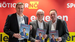 Drei Männer lächeln und posieren für ein Foto, während sie ein Buch mit einem Bild eines Mannes im Anzug halten. Der Mann auf der linken Seite trägt einen Anzug und eine Krawatte. Der Mann in der Mitte trägt eine Brille und einen Anzug. Der Mann auf der rechten Seite trägt einen Anzug. Sie stehen vor einer roten Wand mit gelben und weißen Buchstaben.