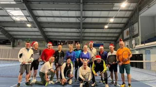 Eine Gruppe von Erwachsenen mit Weihnachtsmützen posiert für ein Foto auf einem Tennisplatz. Einige halten Tennisschläger und andere stehen in der Nähe des Netzes. Der überdachte Platz hat Metallträger und eine Banner an der Wand.