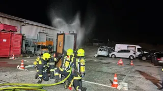 Drei Feuerwehrleute kümmern sich nachts um einen Fahrzeugbrand. Das Fahrzeug steht vor einem Gebäude, und ein Feuerlöscher wird eingesetzt.