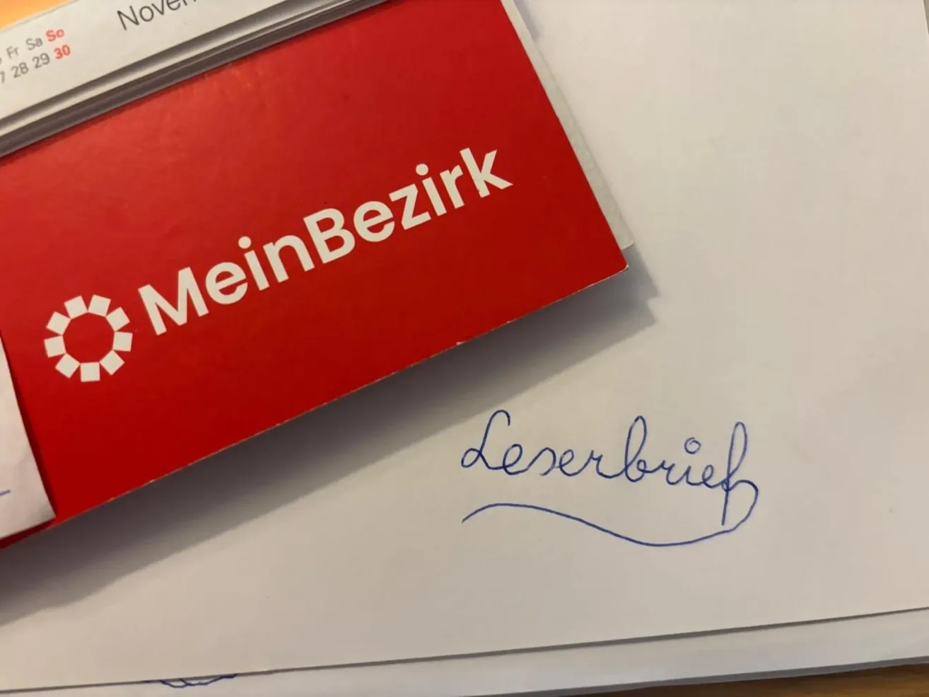 Ein roter Ordner mit der Aufschrift 'MeinBe zirk' liegt auf einem weißen Papier mit blauer kursiver Schrift, die 'Leverbrief' lautet.