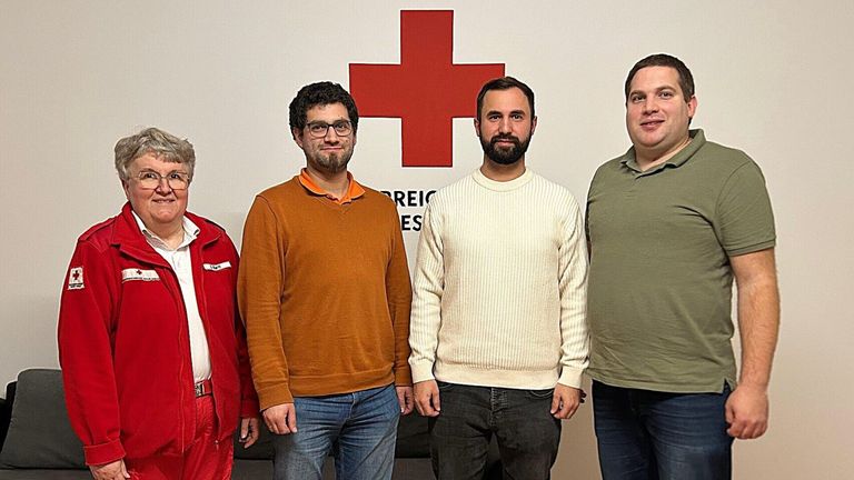 Vier Männer stehen vor einer Wand mit einem roten Kreuz, möglicherweise für ein Foto. Der Mann auf der linken Seite trägt eine rote Jacke.