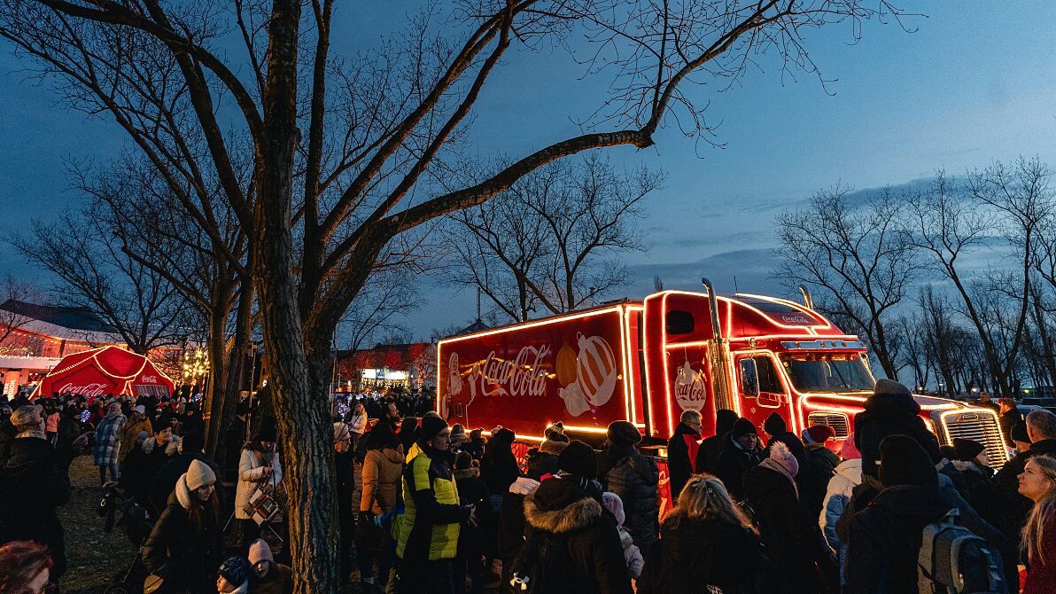 Eine Menschenmenge versammelt sich um einen mit festlichen Lichtern beleuchteten Coca-Cola-Weihnachtslastwagen, der in einem Park bei Dämmerung geparkt ist.