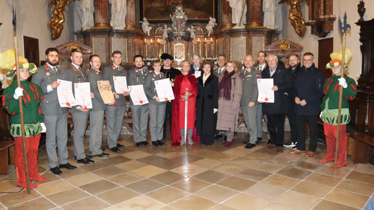 Eine Gruppe von Menschen in Uniformen und mit Urkunden steht in einer Kirche. Eine Frau in einem roten Umhang hält ein Schwert.