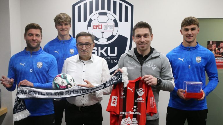 Eine Gruppe von Fußballspielern und Offiziellen hält einen Fußball und ein Trikot. Das Trikot trägt die Aufschrift InterHolzer. Der Mann in der Mitte trägt eine Brille und einen Gürtel.