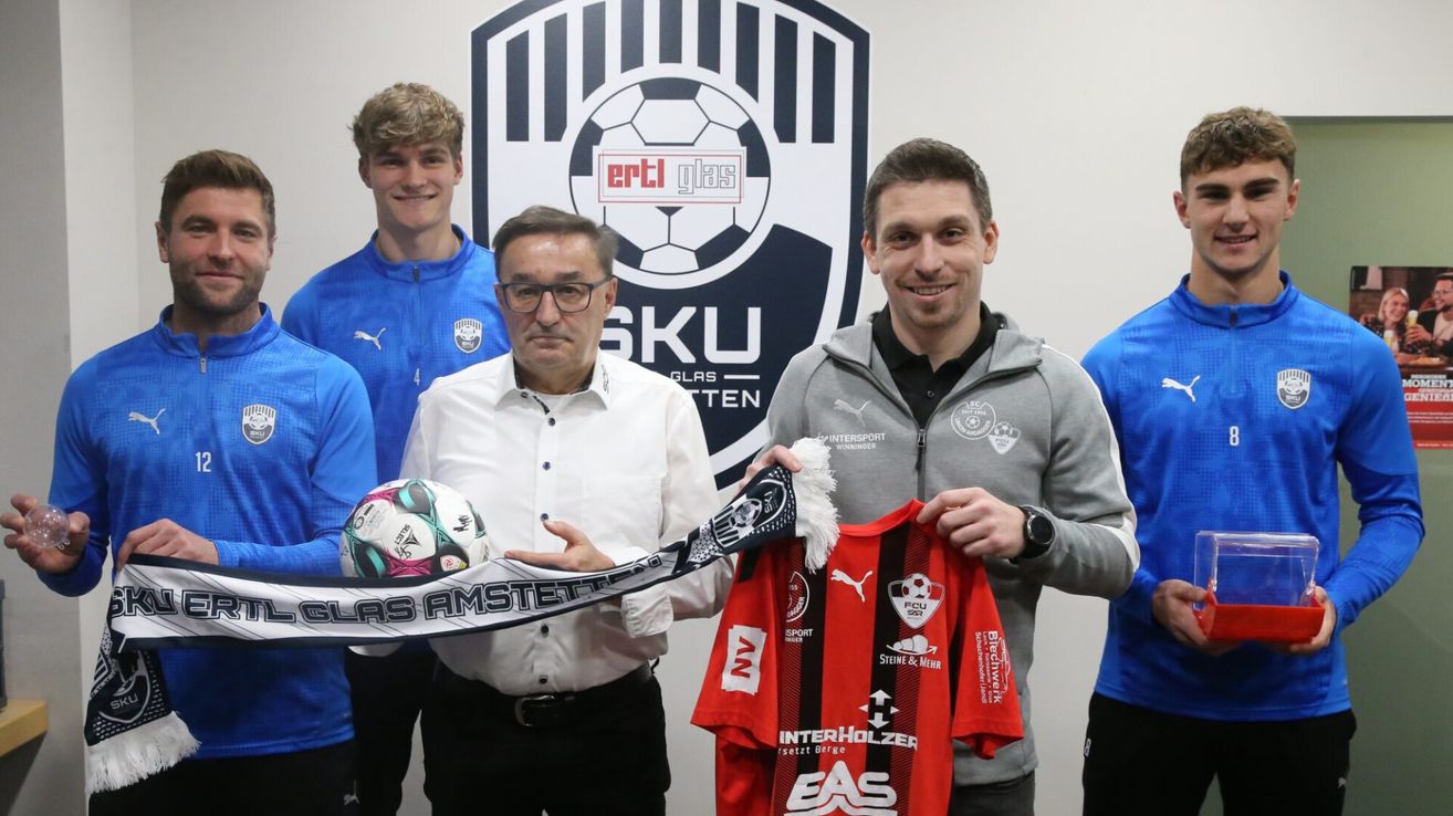 Eine Gruppe von Fußballspielern und Offiziellen hält einen Fußball und ein Trikot. Das Trikot trägt die Aufschrift InterHolzer. Der Mann in der Mitte trägt eine Brille und einen Gürtel.