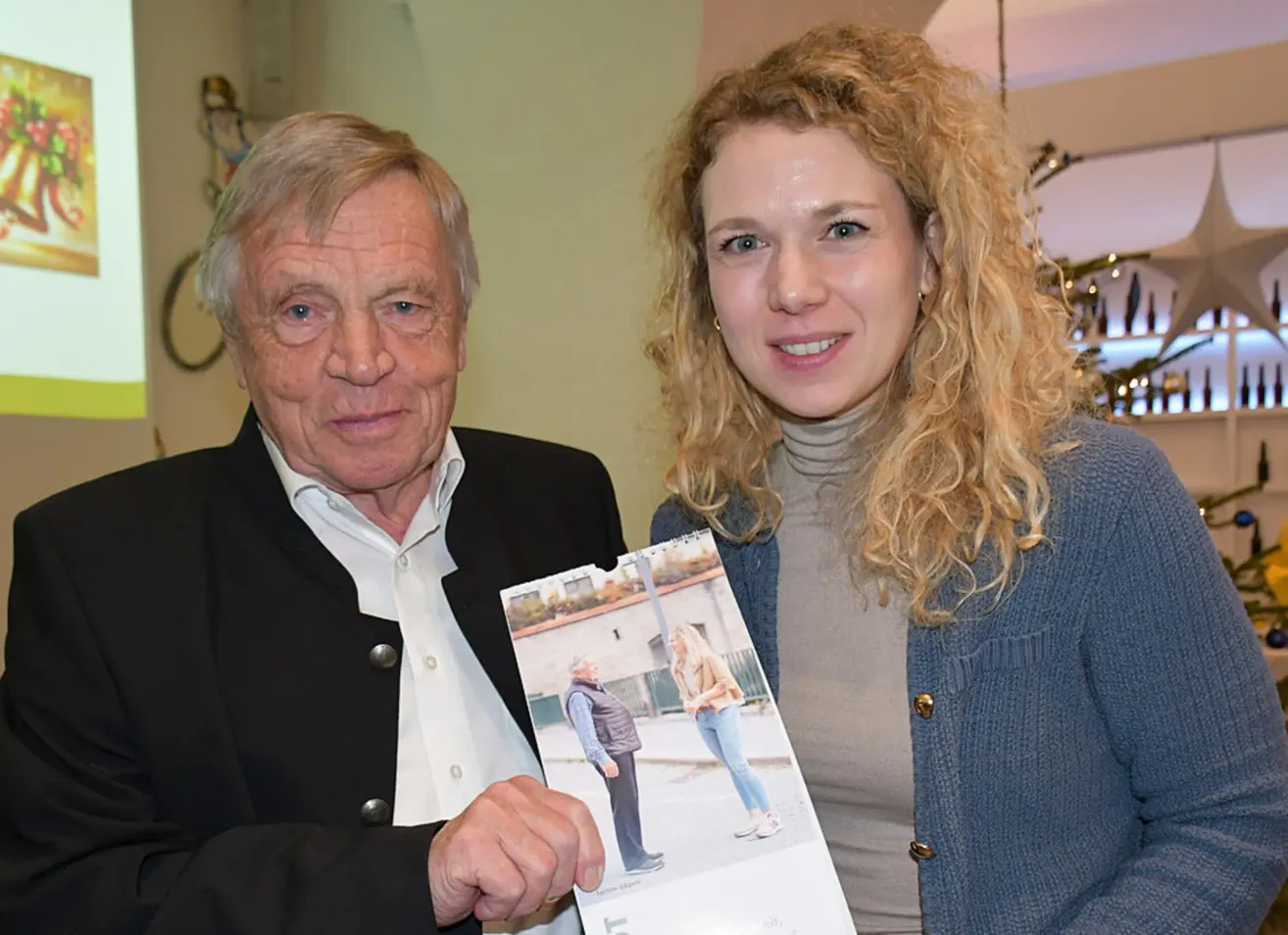 Ein älterer Mann und eine Frau mit blonden Locken lächeln und halten einen Fotokalender. Der Mann trägt einen schwarzen Blazer, und die Frau trägt ein graues Rollkragenshirt und eine blaue Strickjacke.