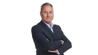 Ein Mann in Anzug steht selbstbewusst mit verschränkten Armen vor schwarzem Hintergrund.