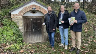 Drei Männer stehen vor einer kleinen, rustikalen Holztür mit einer Ziegelmauer und grüner Vegetation. Sie halten Broschüren und scheinen zu lächeln.