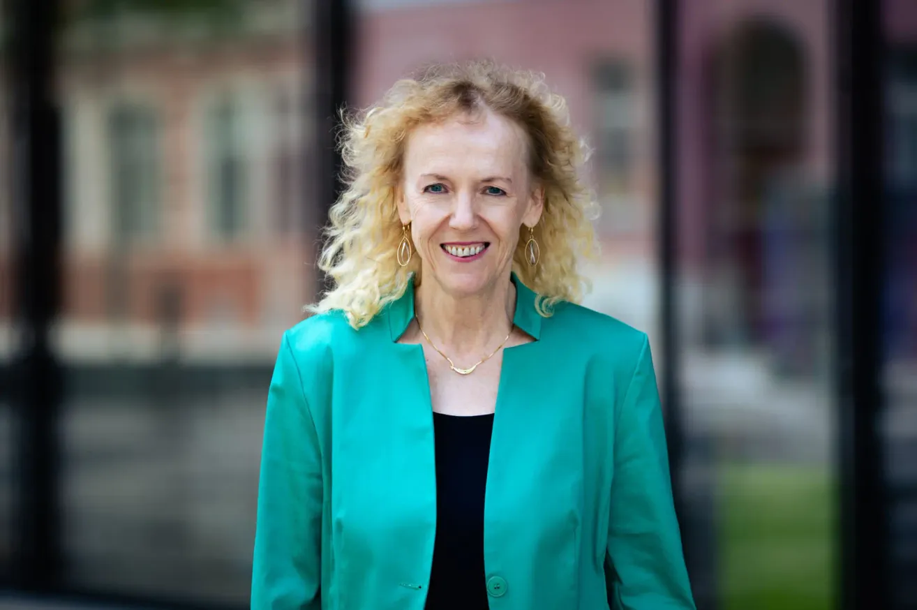 Eine Frau mit lockigem blondem Haar lächelt und steht vor einem Gebäude. Sie trägt einen grünen Blazer und ein schwarzes Top.