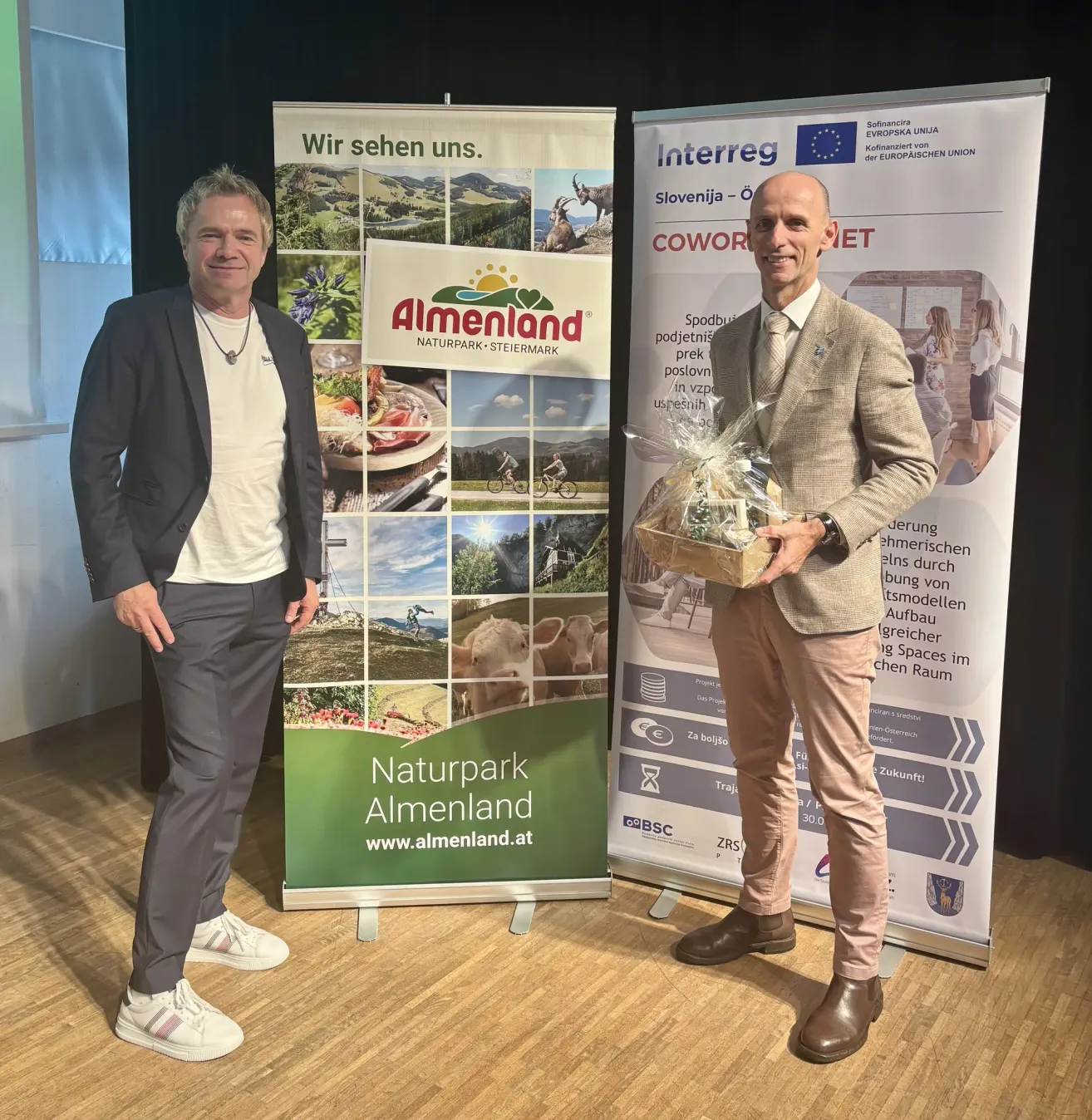 Zwei Männer stehen vor einem Stand mit einem Banner für Naturpark Almenland und halten einen Korb mit Geschenken. Der Mann auf der linken Seite trägt einen Anzug und Sneakers, während der Mann auf der rechten Seite einen Anzug und Lederschuhe trägt.