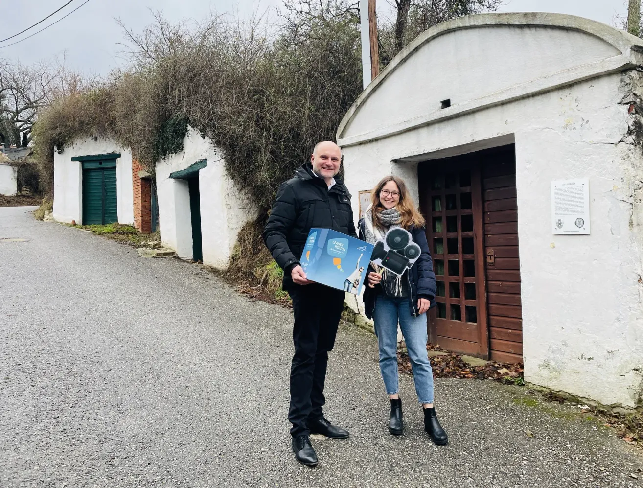 Ein Mann und eine Frau stehen vor einem weißen Gebäude mit einem Poster und einer Kamera in der Hand.