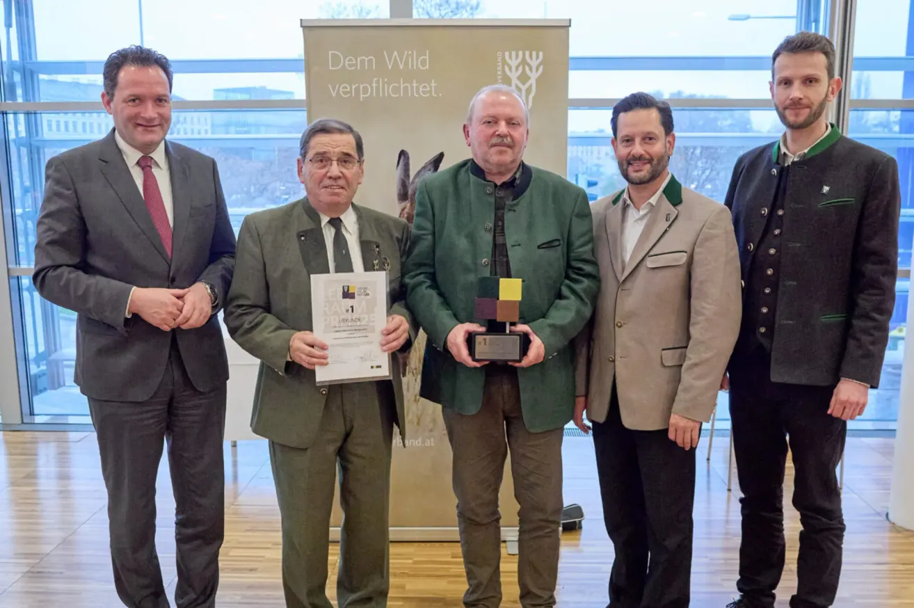 Fünf Männer in formeller Kleidung stehen vor einem Banner. Die beiden in der Mitte halten Auszeichnungen und sehen zufrieden aus. Hinter ihnen ist ein Fenster mit Blick auf ein Gebäude und Bäume.