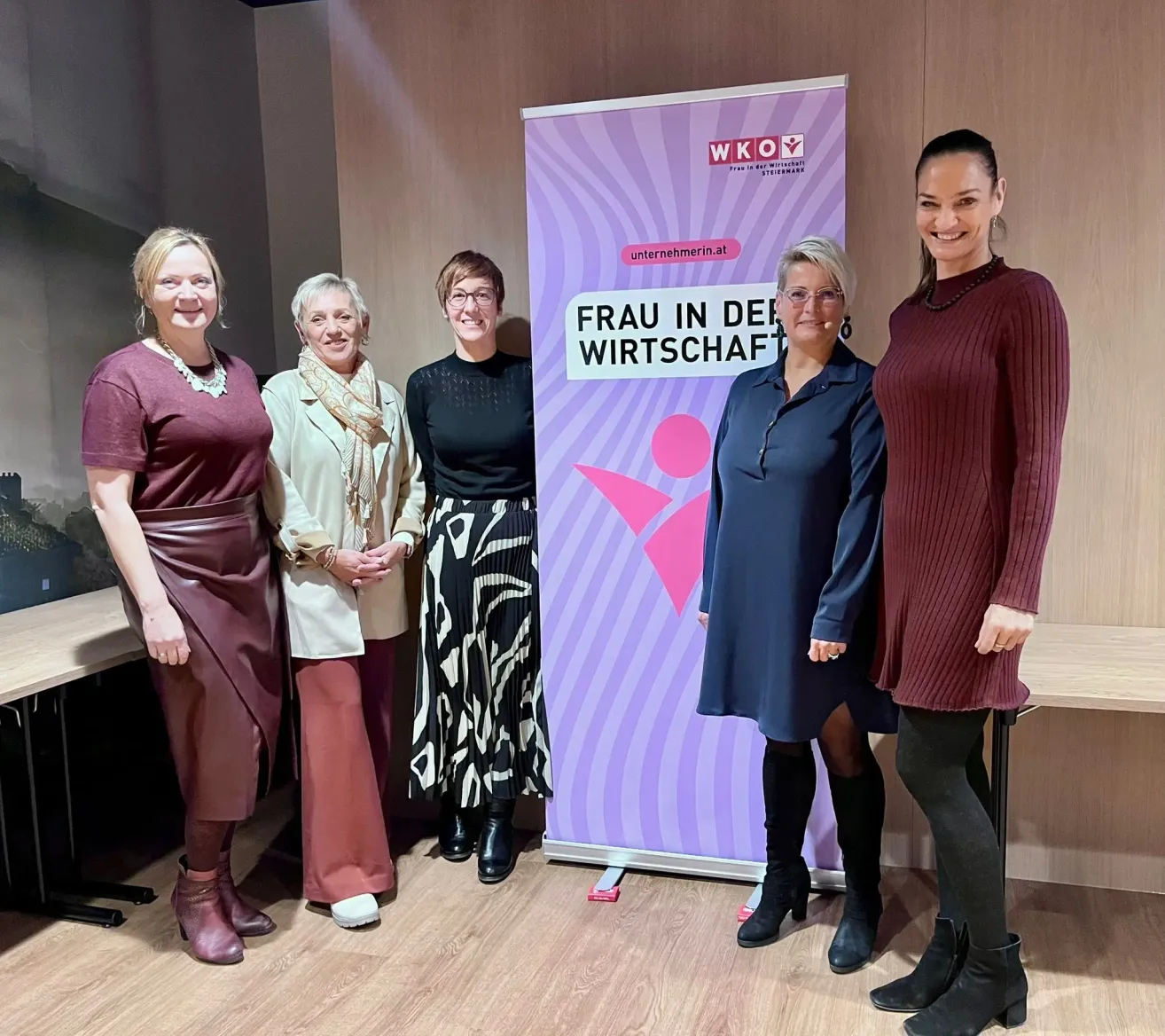 Fünf Frauen stehen neben einem Banner mit der Aufschrift 'Frau in der Wirtschaft'. Sie alle lächeln und scheinen für ein Foto zu posieren. Hinter ihnen steht ein Holztisch.