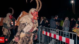 Eine Person in einem roten Teufelskostüm mit Hörnern und einem Korb auf dem Rücken steht vor einer Menschenmenge bei Nacht.