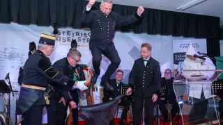 Ein Mann springt in einen Hut, der von zwei anderen gehalten wird, umgeben von anderen in Uniform mit Musikinstrumenten im Hintergrund.