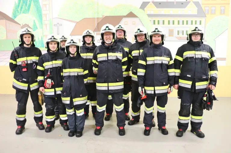 Eine Gruppe von Feuerwehrleuten, alle in Uniform, steht vor einer Kulisse mit Gebäuden und Grün. Die Männer lächeln für ein Foto.