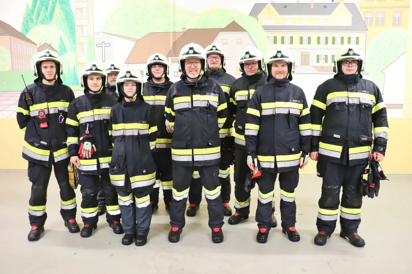 Eine Gruppe von Feuerwehrleuten, alle in Uniform, steht vor einer Kulisse mit Gebäuden und Grün. Die Männer lächeln für ein Foto.
