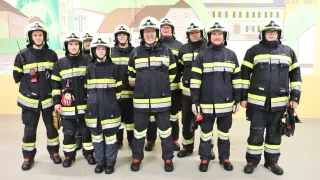 Eine Gruppe von Feuerwehrleuten, alle in Uniform, steht vor einer Kulisse mit Gebäuden und Grün. Die Männer lächeln für ein Foto.