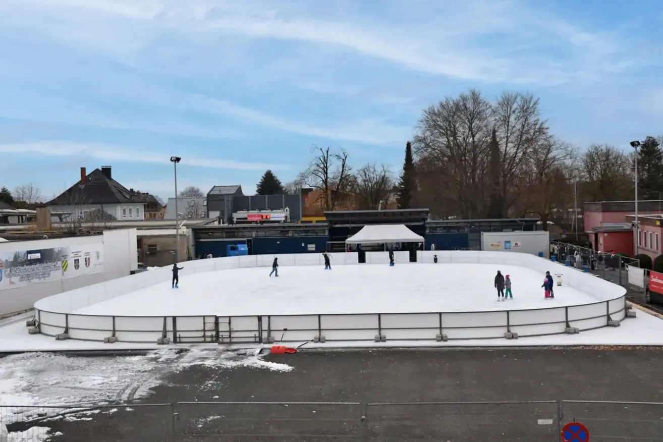 Eine Freiluft-Eisbahn mit mehreren Schlittschuhläufern. Ein Zelt befindet sich rechts. Eine Straßenlaterne steht links. Bäume umgeben die Eisbahn. Gebäude sind im Hintergrund.
