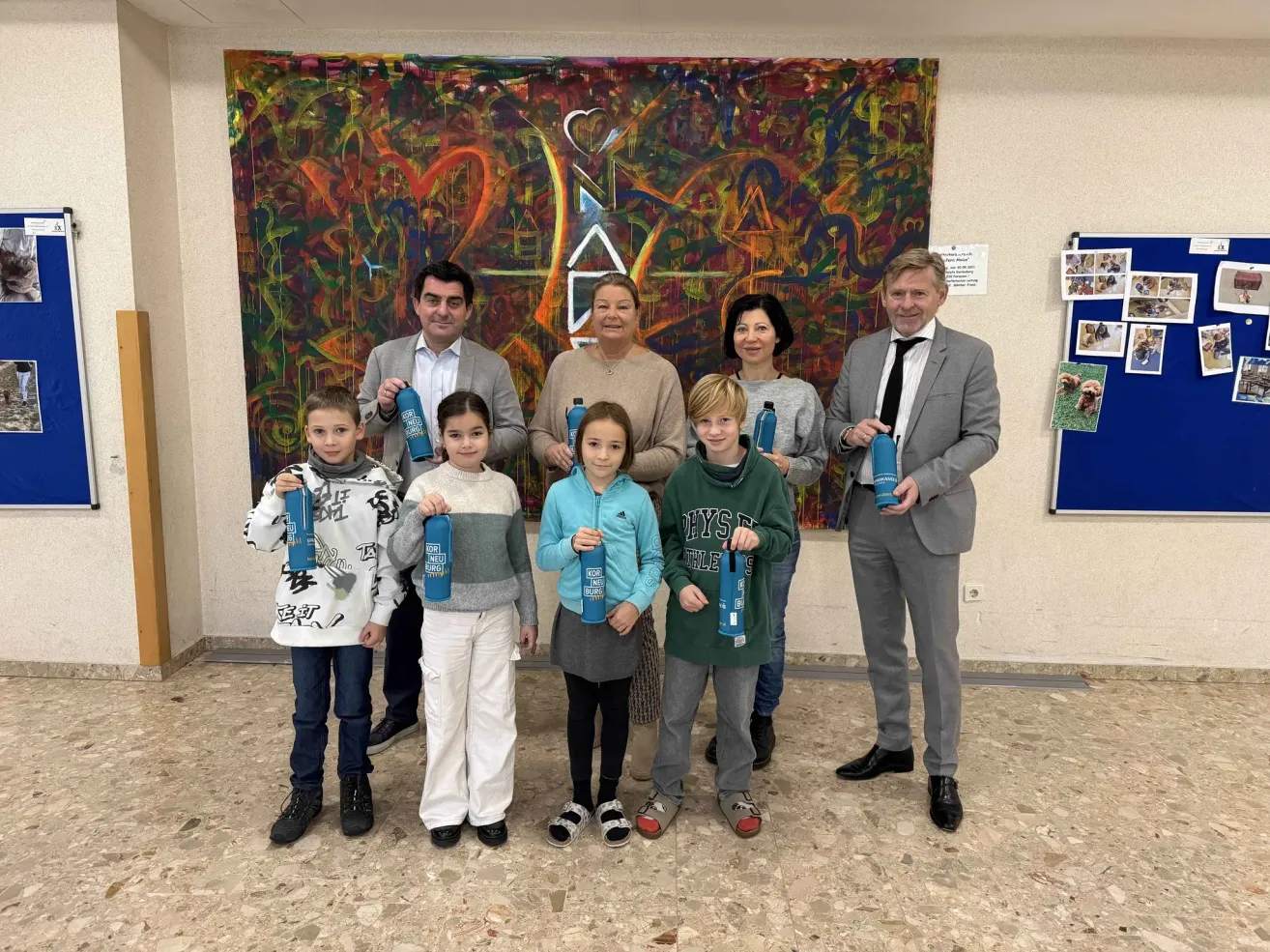 Eine Gruppe von Menschen, einschließlich Erwachsener und Kindern, steht vor einem farbenfrohen Gemälde und hält blaue Flaschen. Sie scheinen für ein Foto zu posieren.