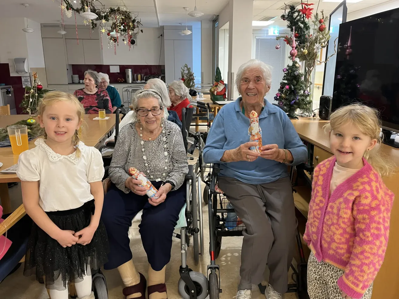 Eine Gruppe älterer Frauen im Rollstuhl posiert für ein Foto mit zwei Kindern. Sie halten Nussknacker und lächeln, mit Weihnachtsdekorationen und einem Weihnachtsbaum im Hintergrund.