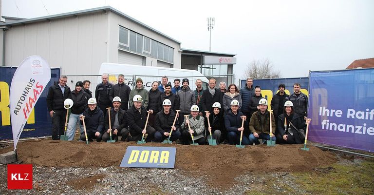 Eine Gruppe von Menschen in Winterkleidung posiert für ein Foto vor einem Gebäude mit einem Schild, auf dem PORR steht. Sie halten Schaufeln in den Händen, und der Boden ist mit Erde bedeckt.