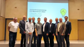 Acht Männer in Anzügen und Krawatten stehen vor einer Leinwand mit einer Präsentationsfolie über eine Generalversammlung.