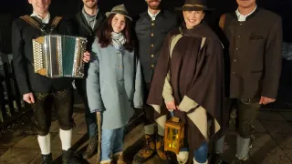 Eine Gruppe von Personen in traditioneller Kleidung posiert für ein Foto, zwei Frauen halten ein Akkordeon und eine Laterne, und ein Mann neben ihnen trägt einen Hut.