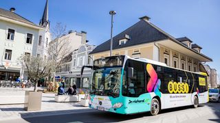 Ein türkisfarbener Bus fährt auf einer Straße an einem Gebäude vorbei, mit Menschen, die auf Bänken im Hintergrund sitzen.